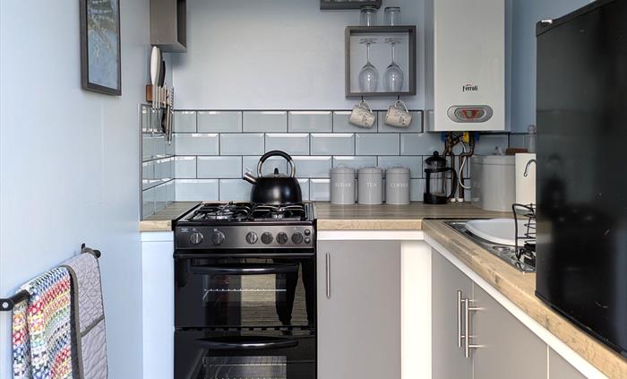 A gas stove with hob top kettle, boiler, sink and black counter top fridge. Tea,coffee,sugar canisters, coffee pot are on the work top.